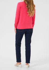 Vrouw met lang golvend haar, gekleed in een felroze top met lange mouwen, donkerblauwe jeans en witte sandalen met hak, staand tegen een effen achtergrond.