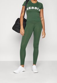 Conjunto de ropa deportiva verde que incluye una camiseta ajustada con el logo "NEBBIA" y leggings de cintura alta a juego. Zapatillas deportivas blancas y una bolsa de malla negra.
