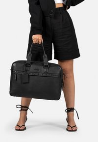 Malette en cuir noir avec doubles poignées et fermetures éclair, dotée d'un logo de la marque en relief et d'une étiquette amovible. Texture lisse, forme rectangulaire.