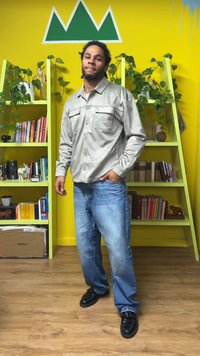 Camicia grigia chiara con bottoni e due tasche sul petto, jeans blu e scarpe nere. L'ambiente presenta scaffali verdi e una parete gialla.