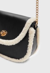 Svart läderhandväska med en texturerad krämfärgad fleecekant, guldfärgade detaljer och en kedjestrapsrem. Har en böjd silhuett och klaffstängning.