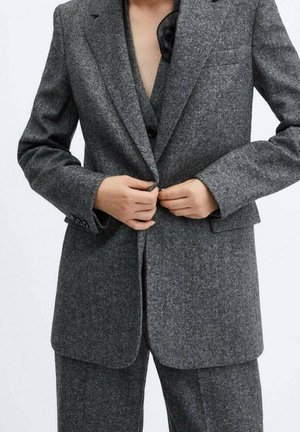 Osoba zapinająca ciemnoszary wełniany żakiet na jeden guzik, z dopasowanymi spodniami, w stylizacji z czarnym topem, na jasnym, jednolitym tle.