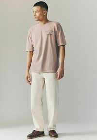 Rundhalset, kortærmet, lyse lilla t-shirt med et trykt logo, parret med hvide bukser og brune snøresko. Glat tekstur.
