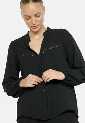 Woman with tied-back hair buttoning a black long-sleeve blouse with lace details across the chest against a white background.