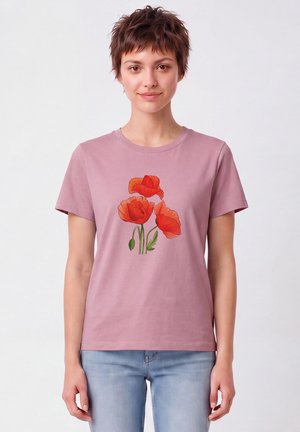 Jeune femme aux cheveux bruns courts portant un t-shirt violet clair avec un motif de coquelicot rouge et un jean bleu clair.