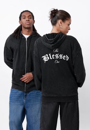 Black zip-up hoodie with front pockets and a textured finish. Back features white script: "The Blessed One." Paired with denim and dark pants.