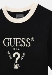 Černé tričko s krémově vroubkovaným výstřihem, se značkovým názvem "GUESS" vyšitým bílou barvou, grafickým motivem otazníku a textem "WASHED JEANS" pod ním.