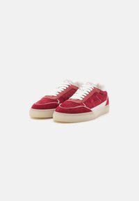 Paire de baskets en daim rouge avec lacets blancs, détail latéral blanc et semelles en caoutchouc beige sur fond clair.