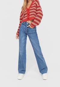 Un pull court à motifs zigzag rouges et crème avec des manches longues, associé à un jean bleu à taille haute et jambes larges, et des baskets blanches.