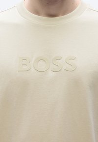 Világos bézs rövid ujjú póló, középen kiemelkedő, texturált "BOSS" logóval. Puha, sima anyag, kerek nyakkivágással.