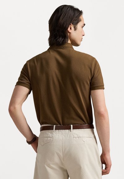Cămașă polo de culoare maronie, cu mânecă scurtă și finisaj texturat, purtată cu pantaloni de culoare deschisă și o curea maronie, fotografiată din spate.