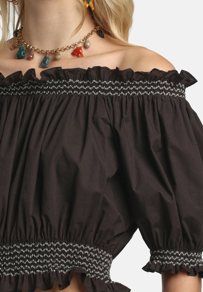 Femme blonde portant un haut noir épaules dénudées avec des bords froncés et un collier chaîne en or avec des pendentifs multicolores en forme d'animaux.