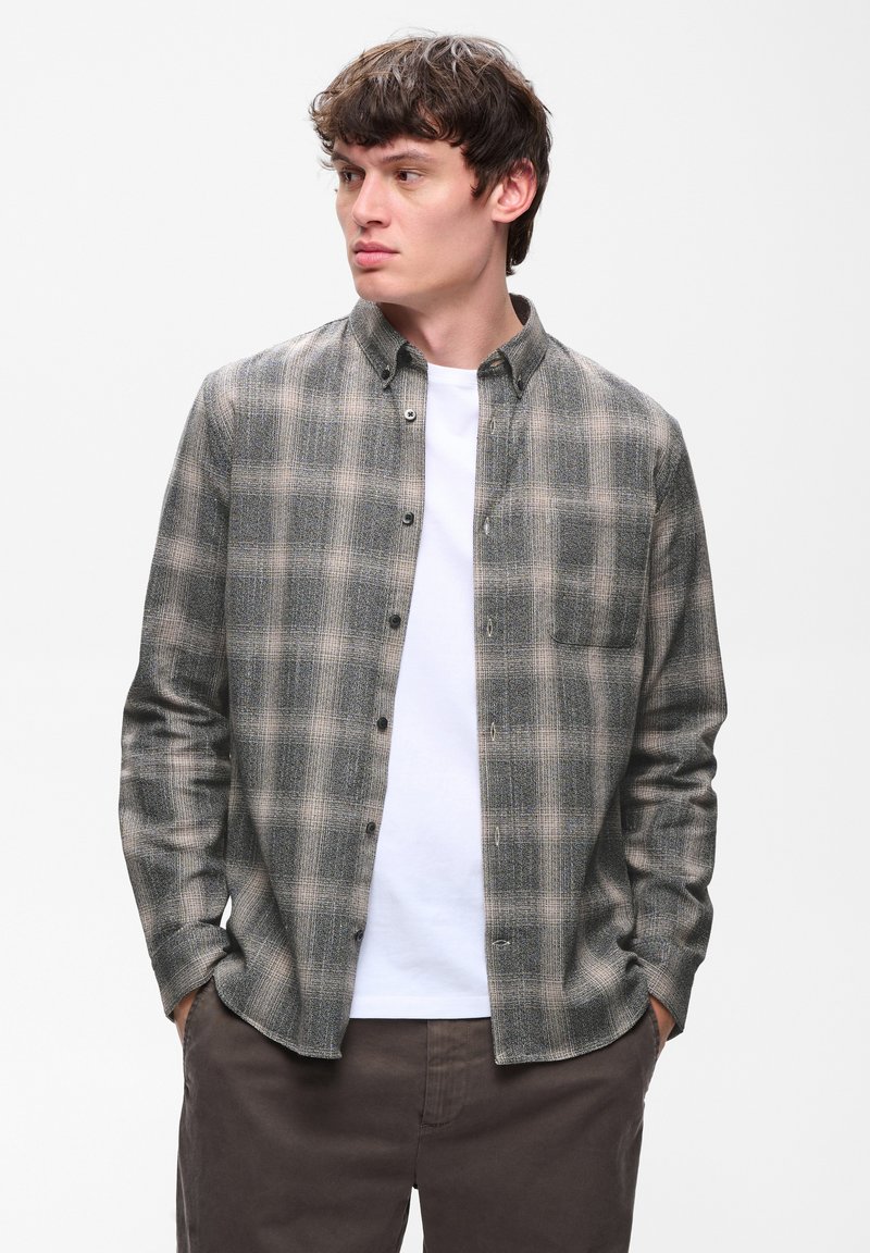 Jeune homme les mains dans les poches, portant une chemise à carreaux grise boutonnée sur un t-shirt blanc et un pantalon foncé, regardant sur le côté.