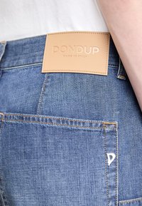 Modré džíny z denimu mají na pasu hnědou koženou nášivku s vyraženým nápisem "DON DUP MADE IN ITALY." Viditelné švy a kapsy.