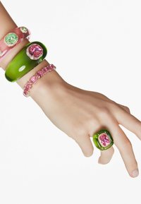 Eine Hand, geschmückt mit mehreren bunten Armreifen, die grüne und pinke Steine aufweisen, sowie einem großen Ring mit einem rosa Stein, vor einem schlichten Hintergrund.