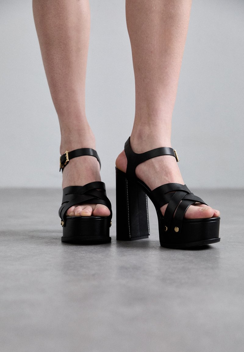 Sandale negre cu platformă și toc masiv, dotate cu bretele încrucișate, cataramă ajustabilă la gleznă și accente din aur.