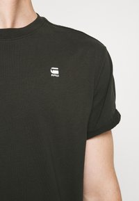 Černé bavlněné tričko s ohrnutými rukávy, na hrudi má malé bílé logo. Hladká textura a klasický design s kulatým výstřihem.