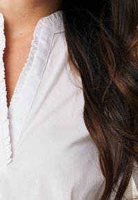 Blouse blanche avec un col froncé et un détail de patte de boutonnage, présentant une texture lisse et un léger éclat, partiellement obscurcie par des cheveux sombres et ondulés.