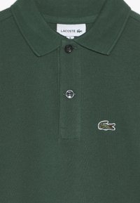 Grön pikétröja i texturerad tyg, med krage, två svarta knappar och en liten broderad krokodillogo på bröstet.