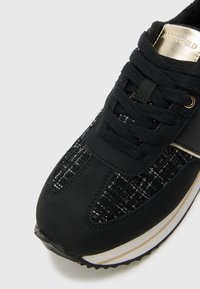 Black sneaker featuring textured fabric with a woven pattern, black laces, and a metallic accent on the heel. Rubber sole with white stripe.