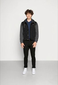 Black denim vest with gray hoodie, navy T-shirt underneath, black tapered pants, and white sneakers. Model stands in a neutral background.