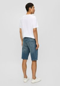 Shorts en denim dans un délavage bleu moyen, avec une coupe droite, des poignets roulés et des poches arrière. Portés avec un t-shirt blanc uni et des baskets blanches.