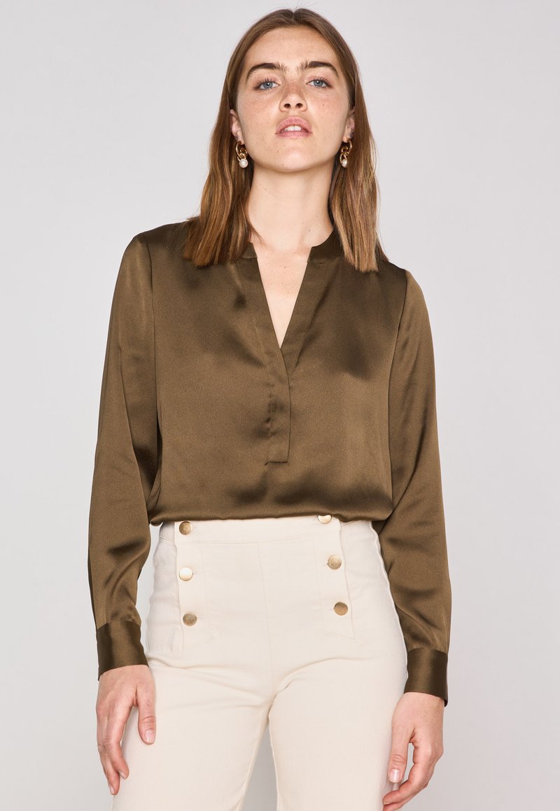 Blusa di raso verde oliva con scollatura a V, maniche lunghe e tessuto uniforme. Indossata con pantaloni a vita alta color crema con bottoni dorati.