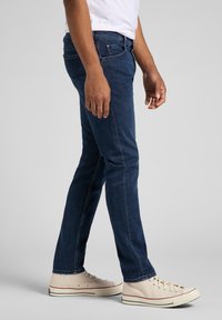 Hombre vestido con jeans ajustados de color azul oscuro y zapatillas de lona color blanco roto, de pie frente a un fondo gris claro liso.