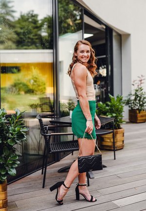 Green textured mini skirt with fringed hem, beige tank top, black heeled sandals, and a black handbag with a chain strap. Outdoor setting.