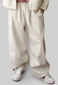 Loose, cream-colored cotton trousers with side pockets, wide legs, and a soft texture, paired with white athletic shoes with accents.