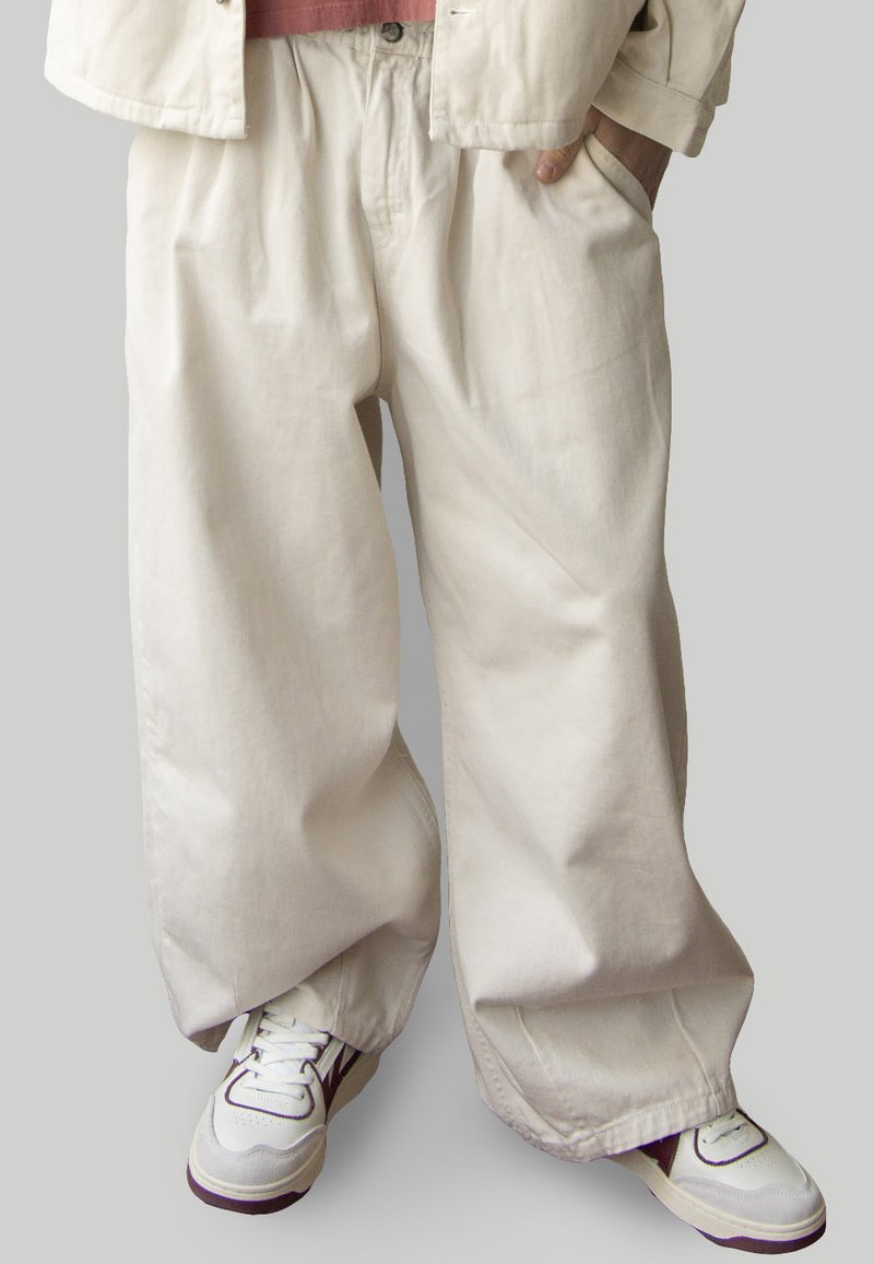 Pantalons en coton lâches de couleur crème avec des poches latérales, des jambes larges et une texture douce, associés à des chaussures de sport blanches avec des accents.