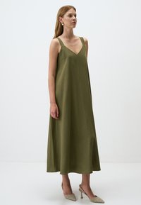 Abito maxi verde oliva con scollatura a V, spalline larghe e silhouette fluida, realizzato in tessuto texturizzato, abbinato a scarpe con tacco metalliche.