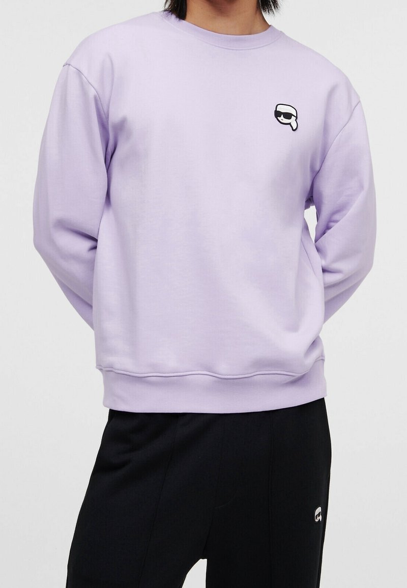 Personne portant un sweat lilas avec un petit logo noir et blanc sur la poitrine et un pantalon noir avec un petit logo assorti.