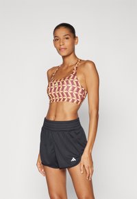Sport bra with a maroon base, featuring a yellow geometric pattern. Paired with black athletic shorts, elastic waistband, and white brand logo.
