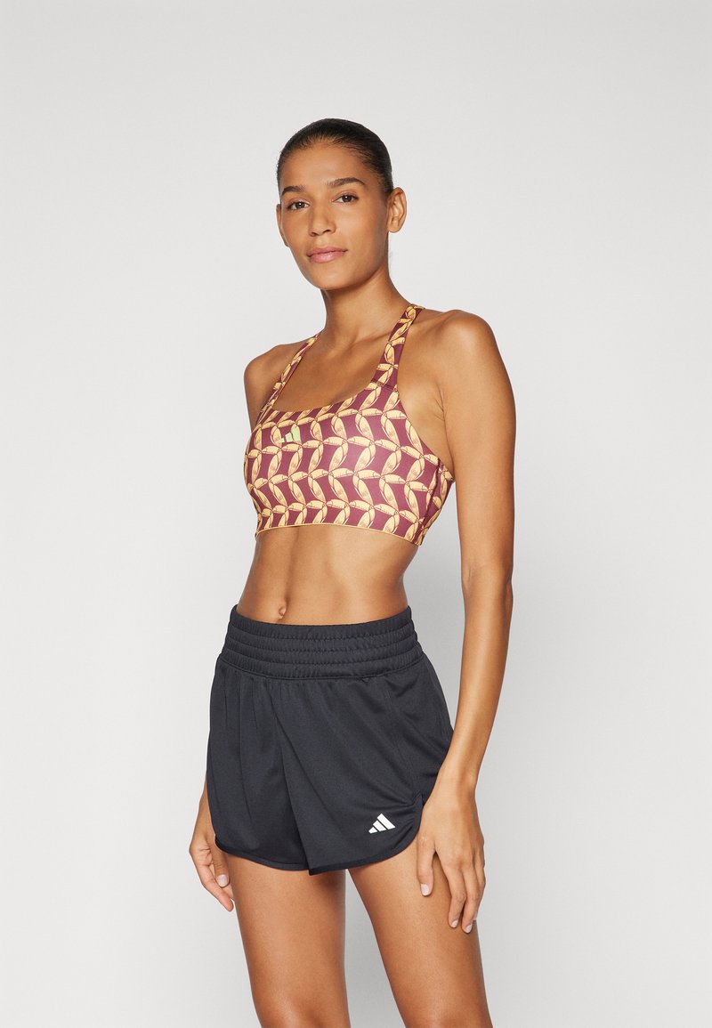 Sport bra with a maroon base, featuring a yellow geometric pattern. Paired with black athletic shorts, elastic waistband, and white brand logo.