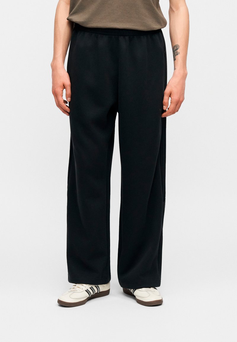 Mand iført løstsiddende sorte bukser, brun t-shirt og hvide sneakers med sorte striber, stående mod en ensfarvet baggrund.
