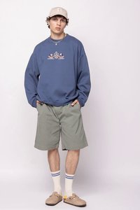 Giovane uomo che indossa un berretto beige, una felpa blu con grafica, pantaloni corti grigi, calze bianche con strisce blu e scarpe slip-on beige, in piedi su uno sfondo neutro.