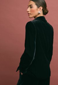 Musträkkidega must samet blazer, millel on sirged õlad ja slim fit lõige, millel on lõhenenud krae ning delikaatsed tasku detailid. Taust on pehme roosa.