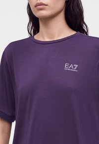 Γυναίκα που φοράει μωβ μπλούζα Emporio Armani EA7 με στρογγυλή λαιμόκοψη και κοντά μανίκια, ορατή από τους ώμους έως το μέσο του στήθους.