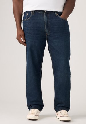 Mężczyzna w ciemnoniebieskich dżinsach o prostych nogawkach, białych trampkach i białej koszuli, stoi z jedną ręką w kieszeni.