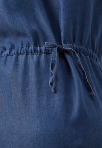 Δυαχτικό ρούχο από τζιν με ζώνη στο μέσο και λεπτομέρεια κορδέλας. Το χρώμα είναι σκούρο μπλε με λεία υφή και διακριτική γυαλάδα.