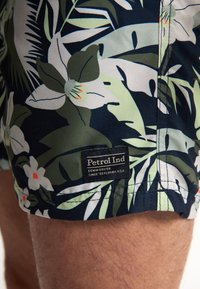 Costumi da bagno con stampa floreale in blu navy, caratterizzati da foglie di palma e fiori bianchi. Include un'etichetta nera con il testo "Petrol Ind".