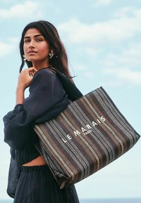 Striped tote bag made from woven material in black, brown, and white. Features "LE MARAIS" printed in white. Sturdy handles, rectangular shape.