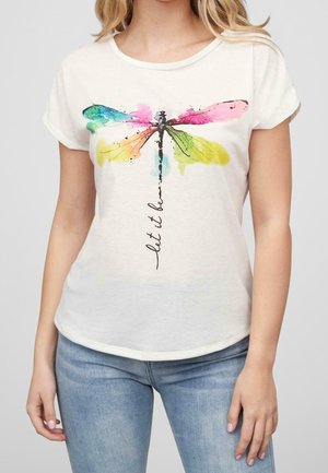 Femme portant un t-shirt blanc avec un motif libellule coloré et le texte "let it be", associé à un jean bleu clair.