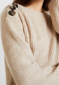 Beige gebreide trui met ribbelstructuur, voorzien van drie grote donkere knopen langs de linker schoudern seam.