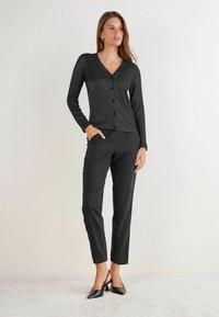 Žena stojící s jednou rukou v kapse, oblečená v černém zapínacím kardiganu, černých kalhotách a černých lodičkách na podpatku s otevřenou patou na světlém pozadí.