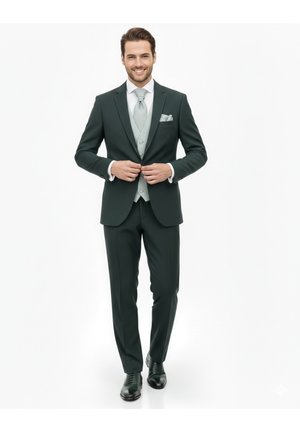 Hombre sonriendo, abotonándose la chaqueta de traje verde oscuro, lleva un chaleco gris claro, corbata, pañuelo en el bolsillo y zapatos de vestir negros sobre fondo blanco.