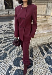 Bordo pritaikytas kostiumas su dvigubu švarku, derančiomis kelnių, ir tekstūruota ruda rankine. Dėvimi bordo aukštakulniai botai.