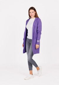 Lange paarse cardigan met twee voorzakken en witte gestreepte manchetten, gecombineerd met een witte top en grijze slim-fit jeans. Witte sneakers.