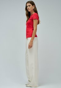 Mujer con cabello largo castaño, usando pendientes de oro, una blusa ajustada de manga corta roja y pantalones anchos blancos, de pie contra un fondo simple.
